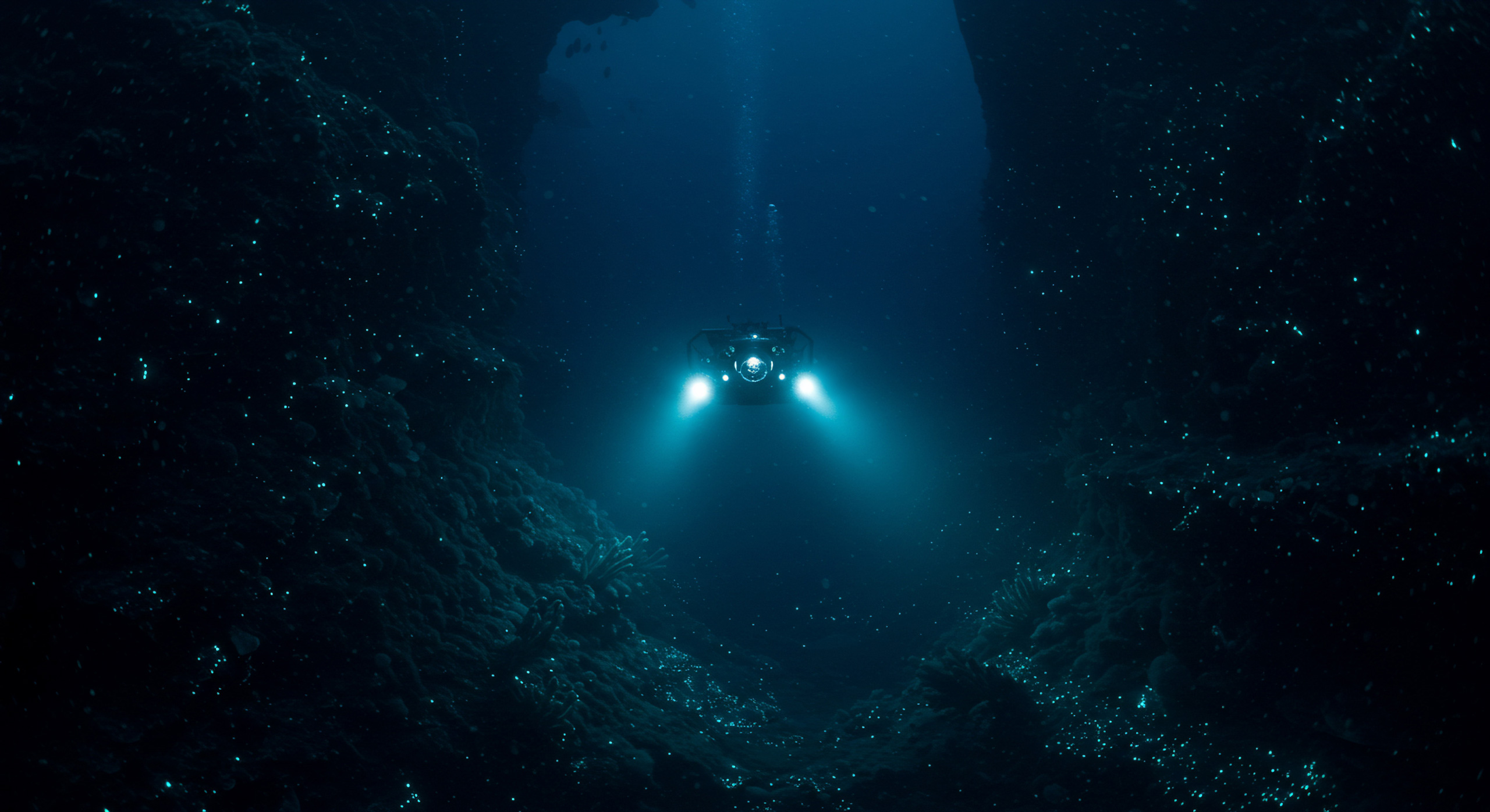 A submersible with bright lights descends into the deep blue ocean with dark rocky surroundings 