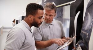 Two machinists reviewing documentation on a clipboard at a CNC machine control panel